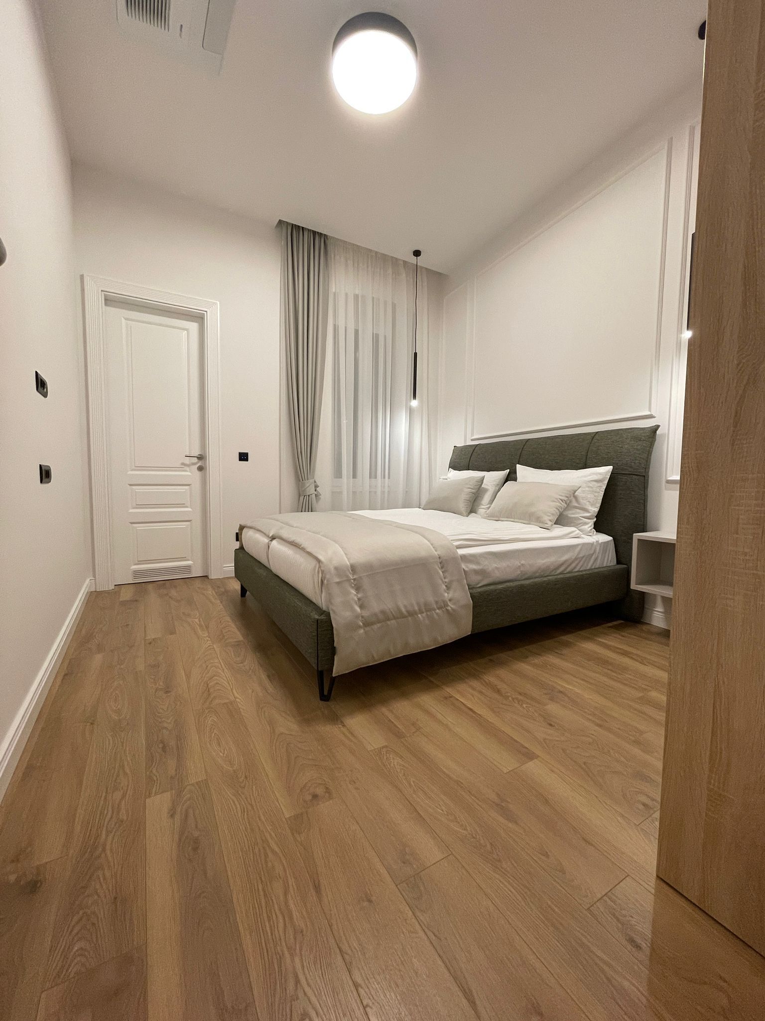 Ein Schlafzimmer mit Bett, Holzboden, einer Tür und einem fenster mit Vorhängen.