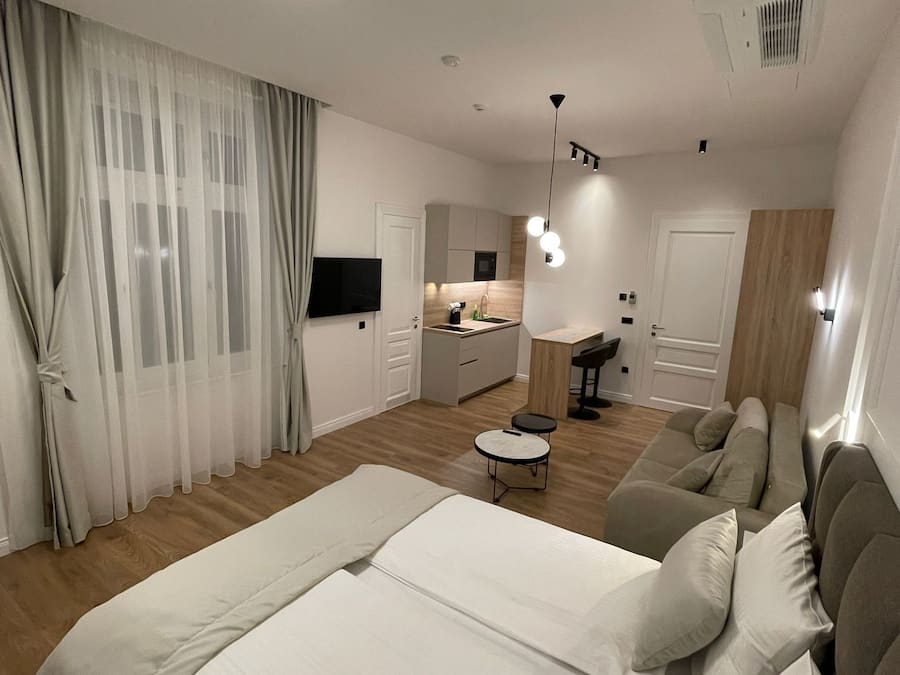 A modern bedroom with a large bed, a sofa, a small table, and a kitchen area in the background.