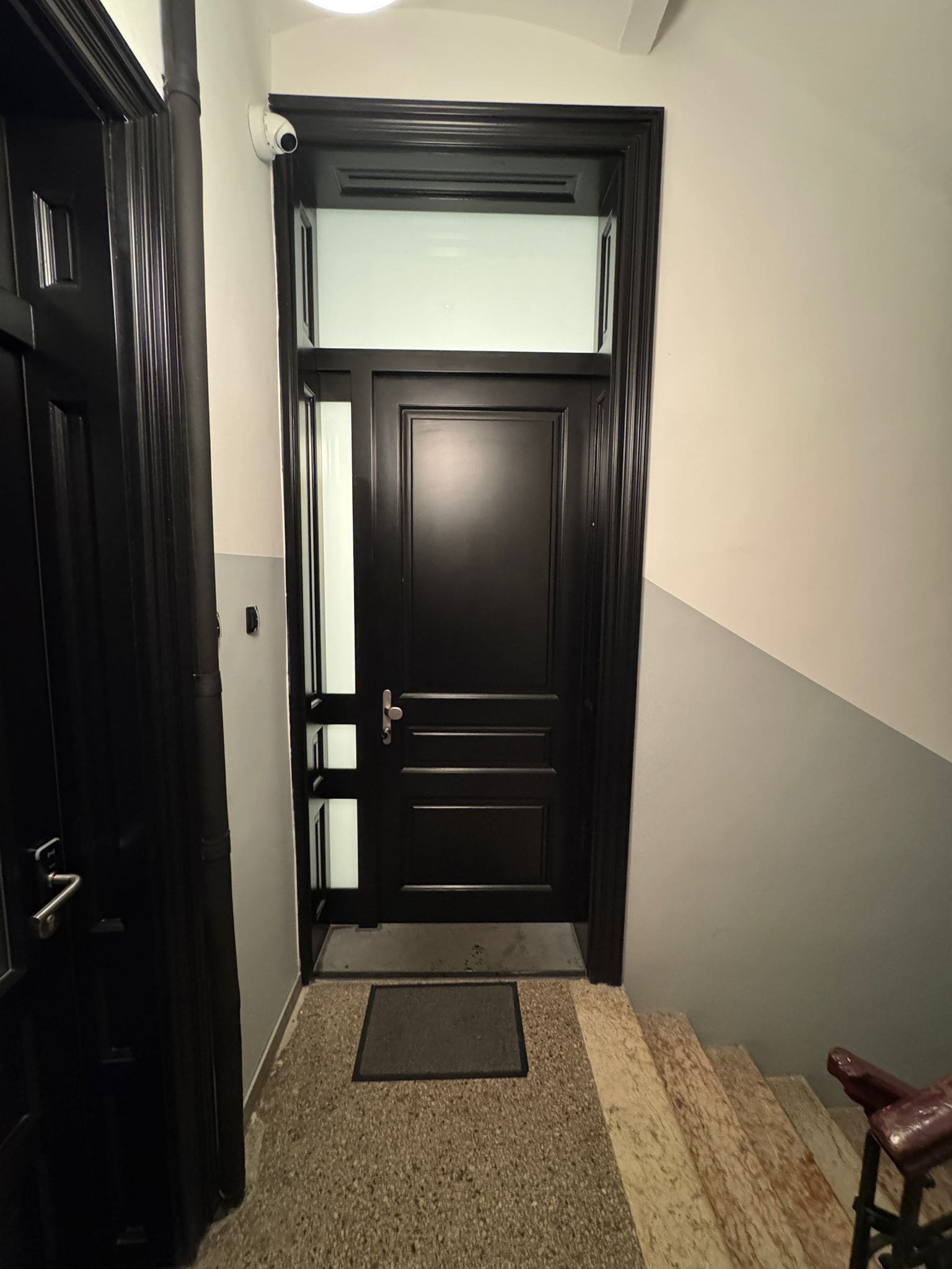 Una puerta de madera oscura con un panel de vidrio satinado, un felpudo y una silla a la derecha.