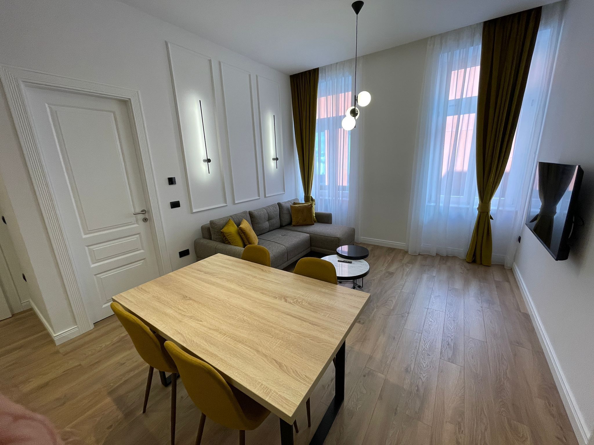 A modern living room with a wooden dining table, yellow chairs, a sofa, and a TV mounted on the wall.