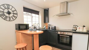 A compact kitchen with a modern design, featuring a black stove, a microwave, and a small refrigerator. There are two wooden stools and a small wooden table. - Well Cottage (Malmesbury)