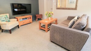 A living room with a flat-screen TV, a wooden coffee table, a sofa, a chair, and a small table with flowers. - Well Cottage (Malmesbury)