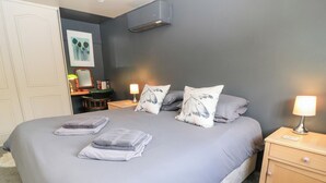 A neatly made bed with a grey bedspread and pillows, a bedside lamp, and a nightstand with a drawer. - Well Cottage (Malmesbury)