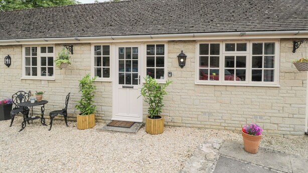 A quaint stone cottage with a tiled roof, a front door, and windows with white frames. There are two potted plants on either side of the door and a small table with chairs in front. - Well Cottage (Malmesbury)