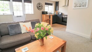 A modern living room with a sofa, a coffee table with a vase of flowers, a kitchen in the background, and a clock on the wall. - Well Cottage (Malmesbury)