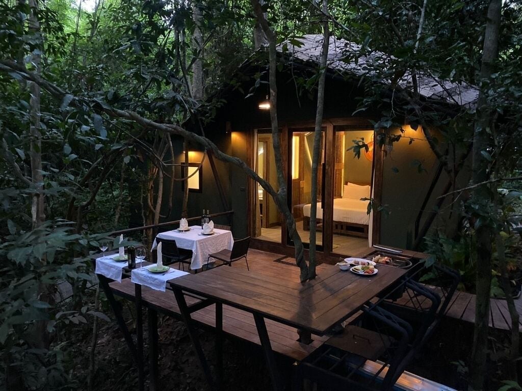 A wooden dining table set with white tablecloths and candles, surrounded by chairs, in a forested area with a building in the background.