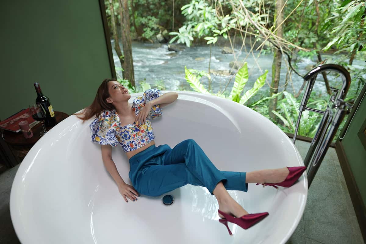 A woman reclining in a large bathtub with a view of a river outside.