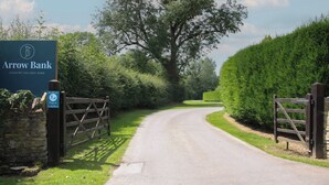 A country holiday park entrance with a sign for Arrow Bank, a paved road, and a wooden gate. - Foxglove (Leominster)
