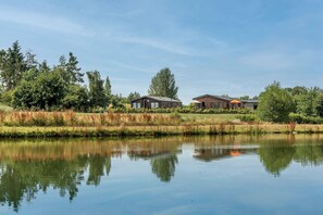 A tranquil lakeside setting with two modern buildings, a clear sky, and a calm water surface reflecting the surroundings. - Foxglove (Leominster)