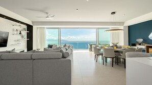 A modern living room with a large gray sectional sofa, a dining area with a table and chairs, and a view of the ocean through large windows. - Stunning Beach Front Condo in Peninsula Tower 1! 3BD Condo for rent in North Hot (Puerto Vallarta)