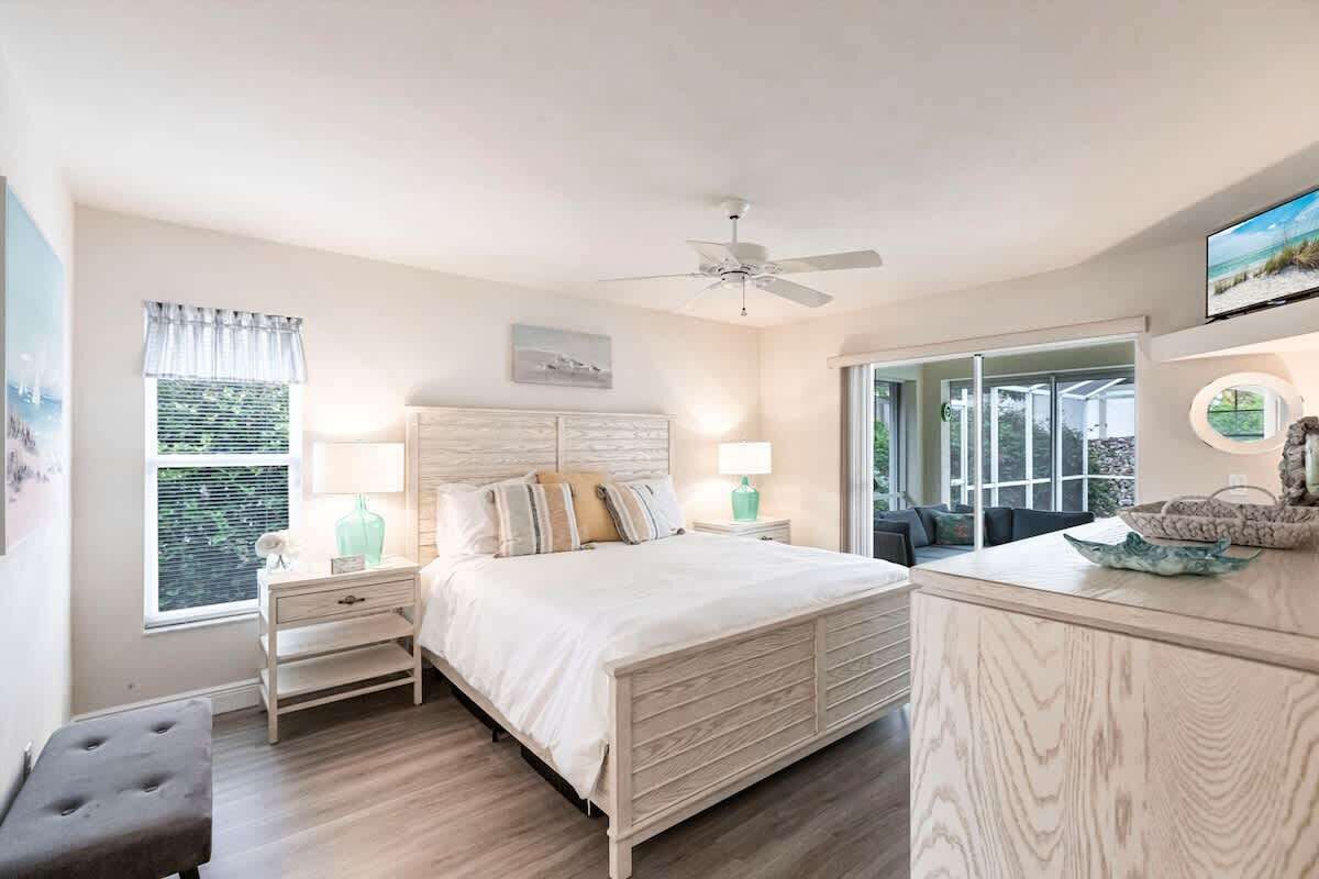 A bedroom with a bed, bedside tables, a nightstand, a bench, a ceiling fan, a window with curtains, a wall-mounted TV, and a view through a glass door.