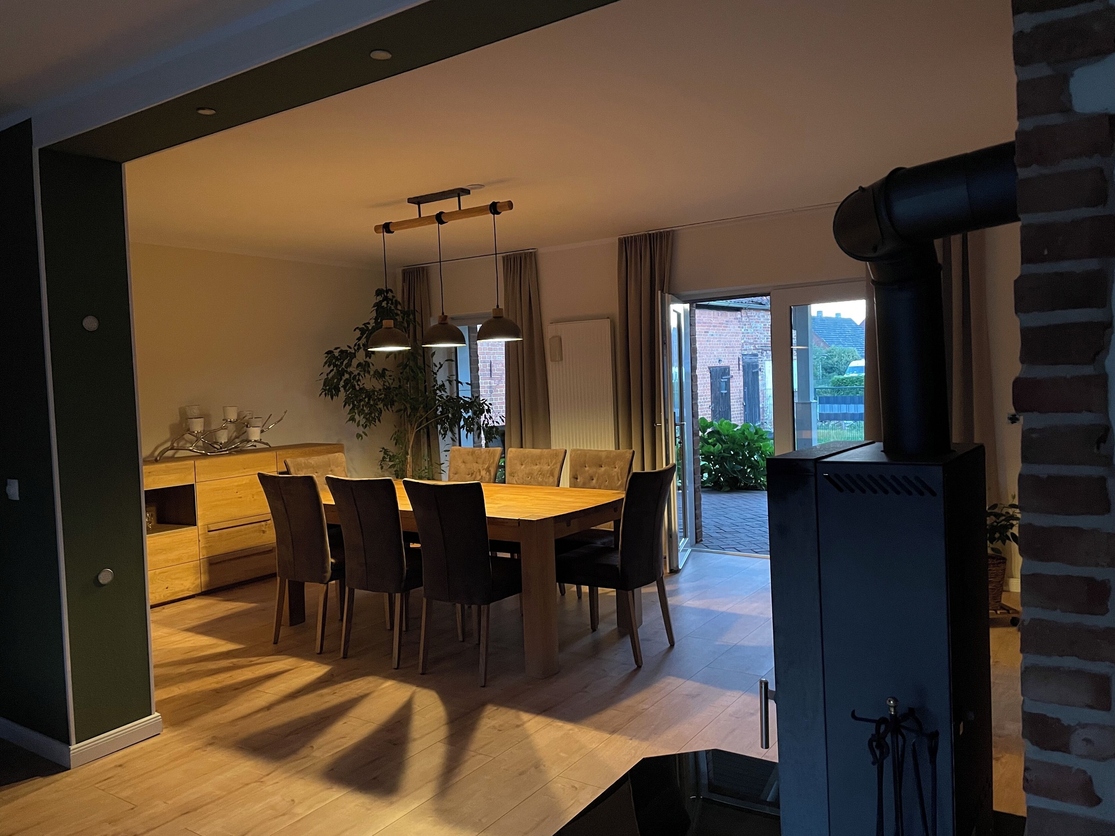 Uma sala de jantar com mesa e cadeiras de madeira, luminárias pendentes, uma planta e vista para o exterior.