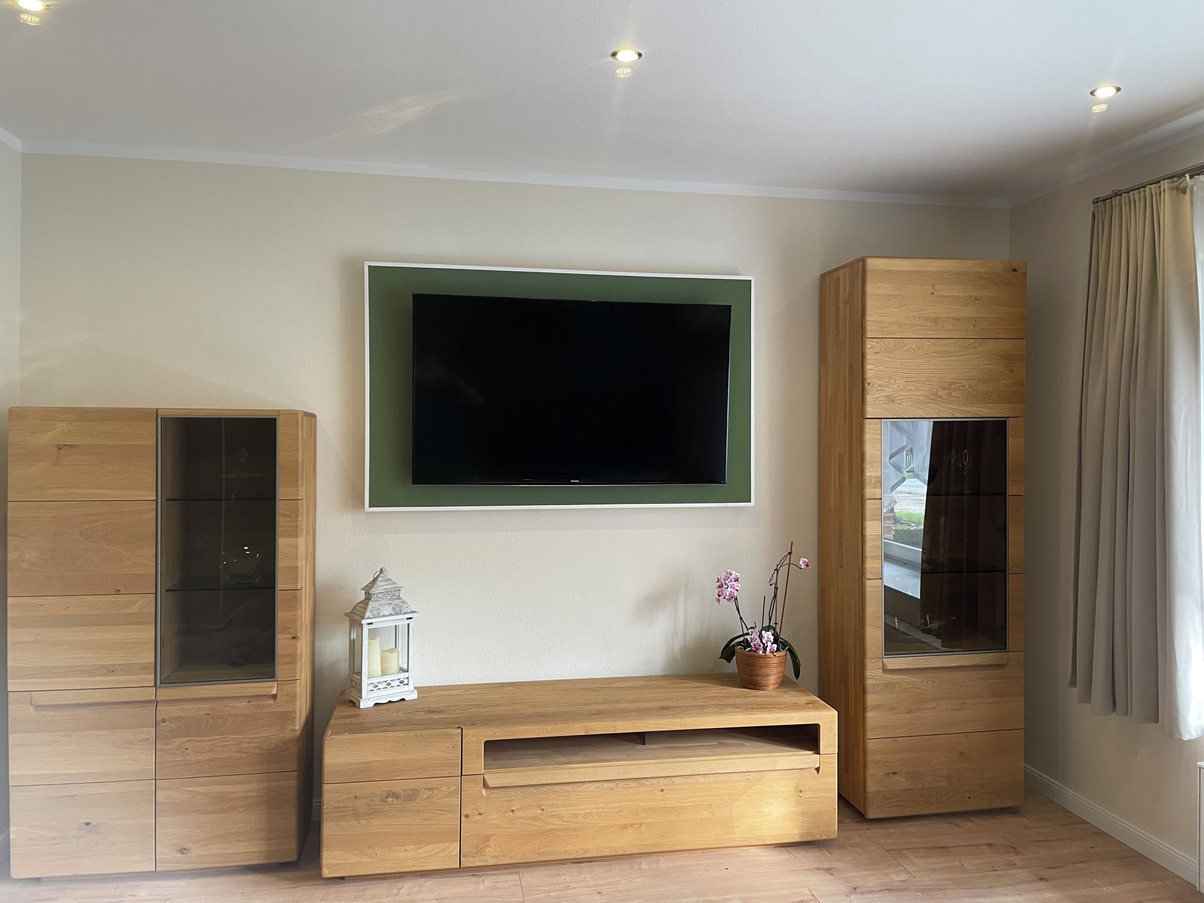 Una sala de estar con un televisor de pantalla plana grande montado en la pared, armarios de madera y una pequeña mesa con un farolillo y una planta en maceta.