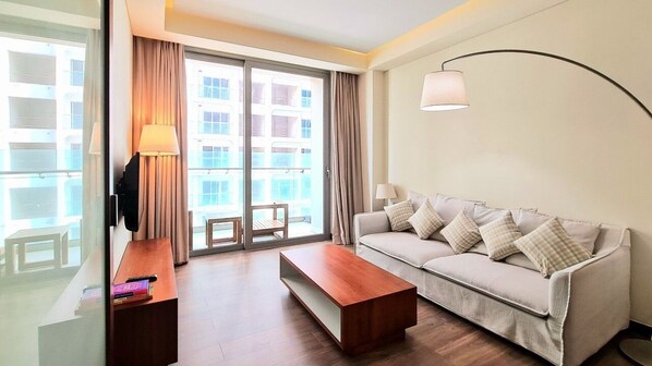 A modern hotel room with a sofa, a wooden coffee table, and a large window with curtains.