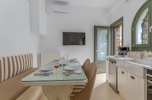 A dining area with a table set for four, a sofa with striped cushions, a television mounted on the wall, and a kitchen with a dishwasher and a window with a view. - Holiday Apartment 'Mrs Kalis Cottage' with Mountain View, Garden & Wi-Fi (ANDROS)