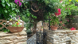 Um caminho de pedra leva a um arco de madeira com um portão de metal, cercado por vegetação exuberante e plantas em vasos.