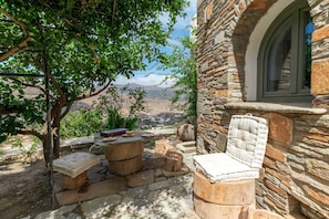 A stone building with a window, a cushioned chair, a small table, and a view of a valley. - Holiday Apartment 'Mrs Kalis Cottage' with Mountain View, Garden & Wi-Fi (ANDROS)