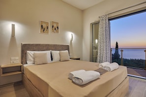 Une chambre d’hôtel avec un grand lit, une table de chevet, des lampes fixées au mur, une vue sur la mer et un balcon avec une porte vitrée.