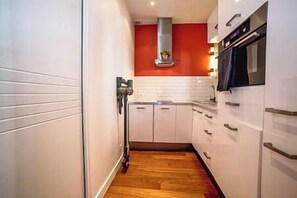 Une cuisine étroite avec des meubles blancs, un plancher en bois et un mur d’accent rouge.