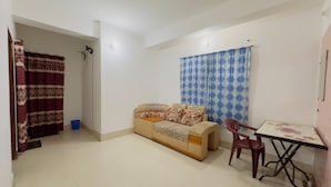 A small, well-lit room with a sofa, a dining table, and a chair. A curtain is partially drawn, and a blue patterned curtain hangs on the wall. - Mohammadia Restaurant & Guest House (Dhaka)