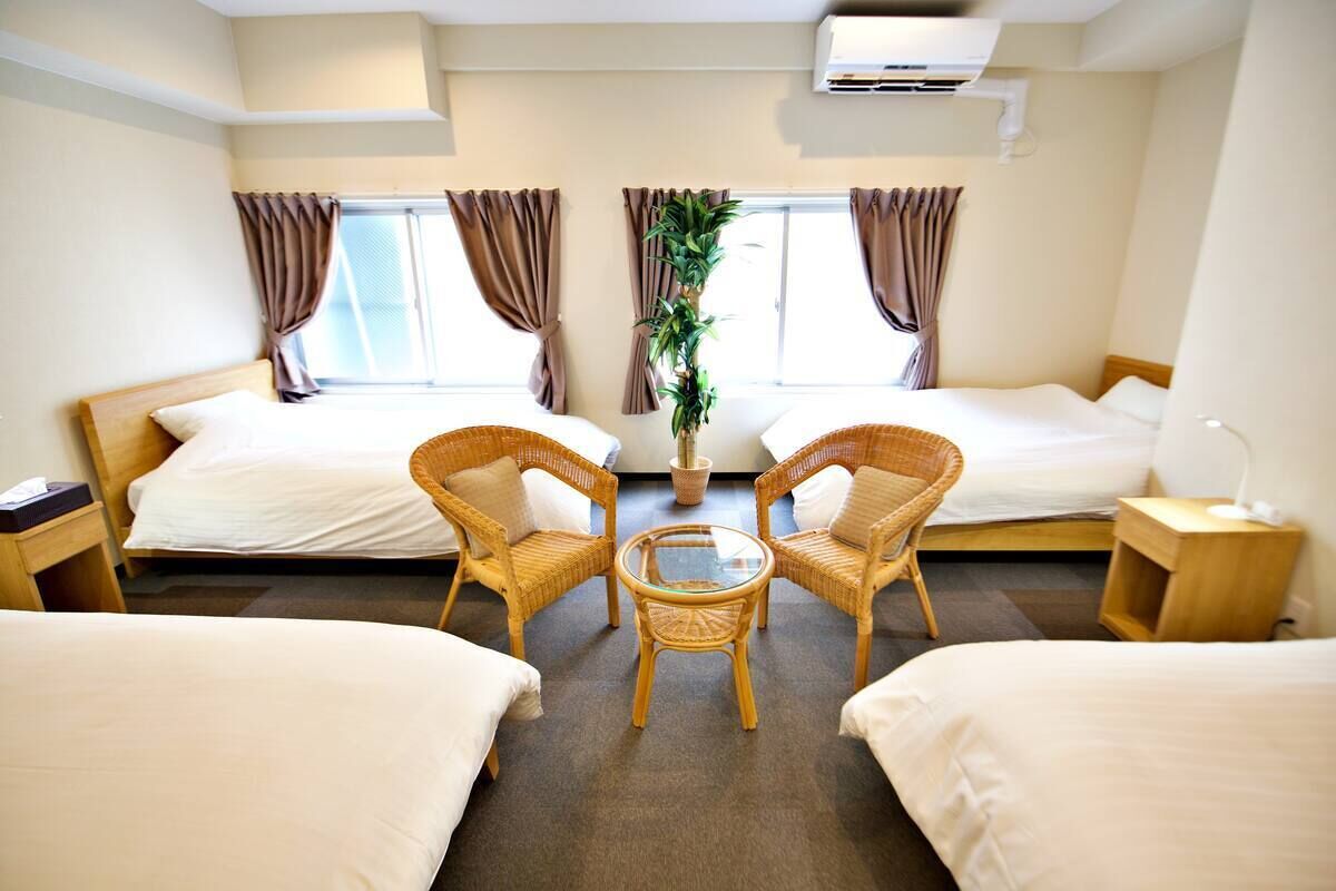 A hotel room with two single beds, a small round table, two wicker chairs, a potted plant, and a nightstand with a lamp.