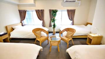 A hotel room with two single beds, a small round table, two wicker chairs, a potted plant, and a nightstand with a lamp.