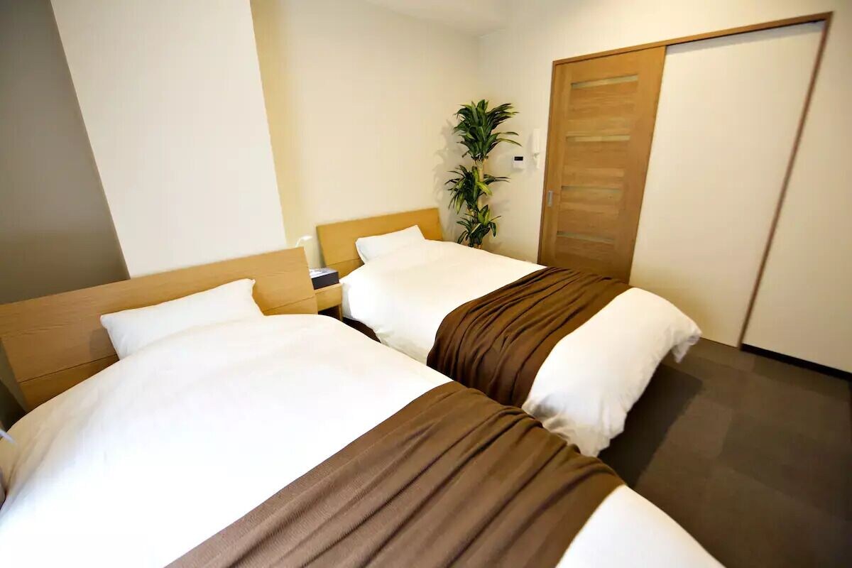 A hotel room with two beds, a wooden headboard, a bedside table, a nightstand, a potted plant, and a sliding door.