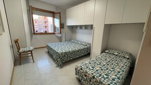 A compact room with two beds, a chair, and a window with a view of buildings. - Alghero Lido sunny beach apartment ☀️ (Alghero)