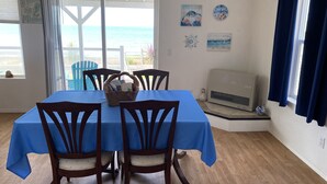 A dining area with a table covered in blue, four wooden chairs, a basket with magazines, a view of the ocean through a glass door, and a wall with a clock and a painting.