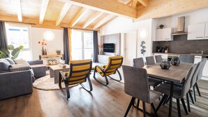 Een moderne woonkamer met een bank, een salontafel en een eethoek met stoelen en een tafel. In de achtergrond bevindt zich een keuken met kastjes en een fornuis.