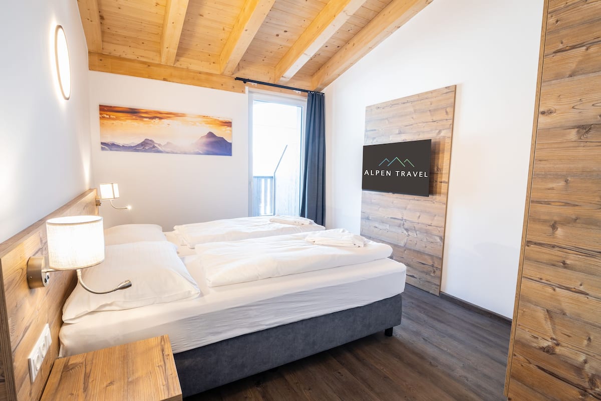 Ein modernes Hotelzimmer mit Holzboden, einem großen Bett, einem Nachttisch mit Lampe, einem Wand-Fernseher und einem Gemälde einer Berglandschaft.