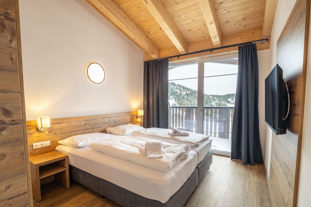 Ein Schlafzimmer mit Holzboden, einem Bett mit weißer Bettwäsche, einem Nachttisch mit Lampe und einem an der Wand befestigten Fernseher.