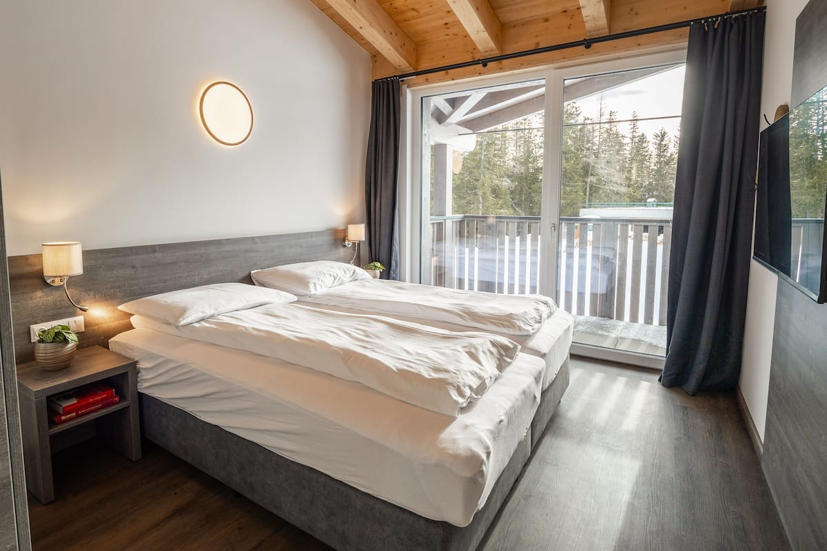 Ein Schlafzimmer mit einem Bett, Nachttischen, einer Lampe, einer Topfpflanze und Blick ins Freie durch ein großes Fenster.