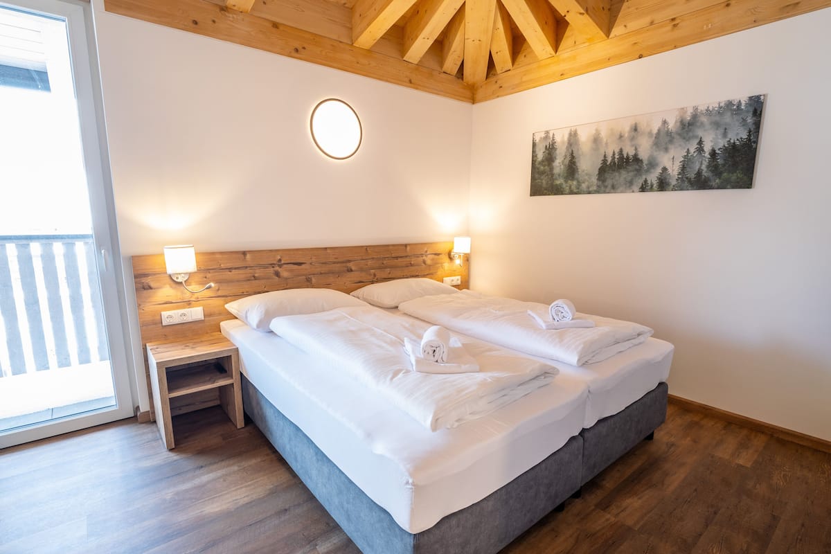 Ein Doppelbett mit weißer Bettwäsche, zwei Nachttische mit Lampen, ein Gemälde eines Waldes und Holzbalken an der Decke.