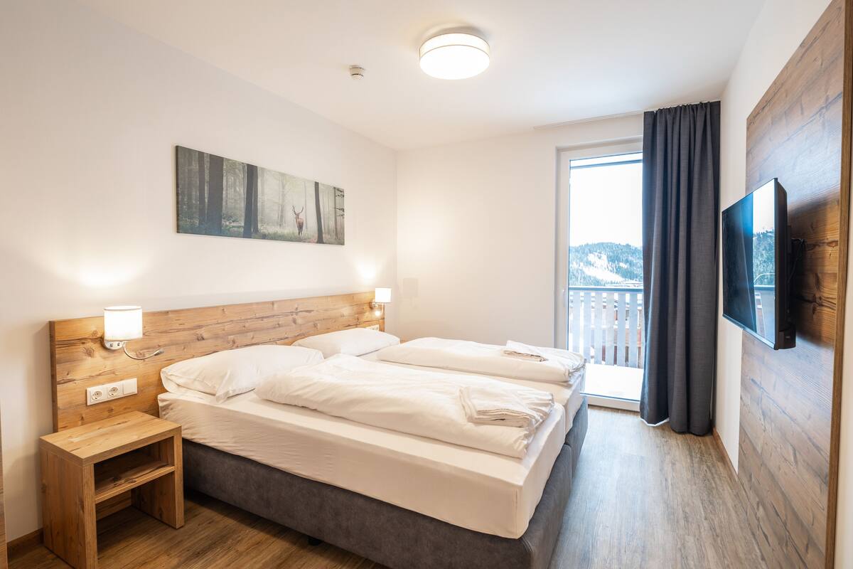Ein Hotelzimmer mit einem hölzernen Kopfteil, zwei Betten, einem Nachttisch, einem Wand-Fernseher und einem Fenster mit Vorhängen.