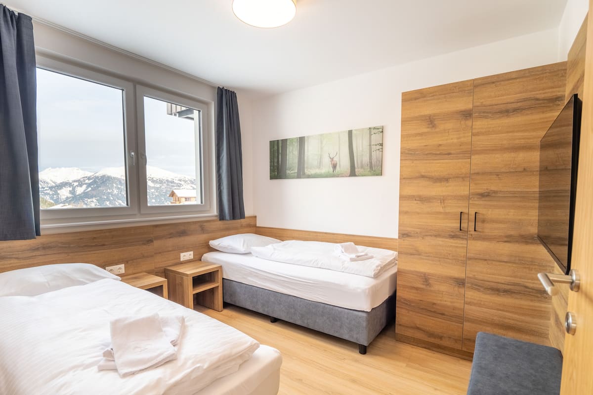Ein Hotelzimmer mit zwei Betten, einem Holzschrank, einem Gemälde und einem Fenster mit Bergblick.