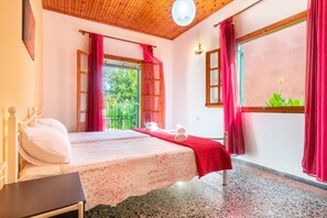 A bedroom with a bed, a bedside table, a window with curtains, and a view of greenery outside.