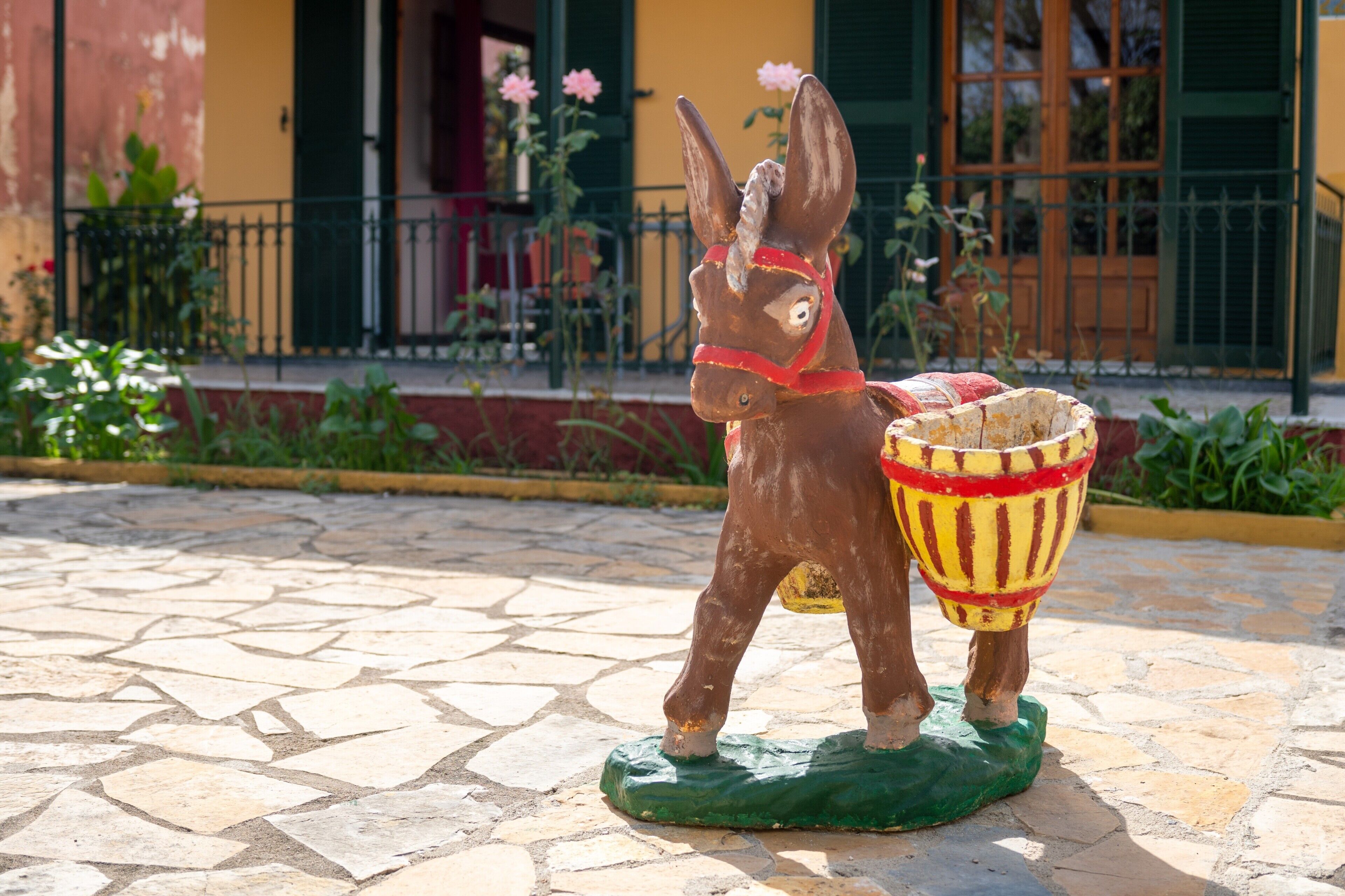A statue of a donkey carrying a basket on a green base, set on a stone-paved area with a building in the background.