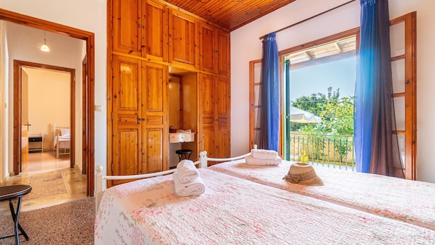 Une chambre avec des armoires en bois, un balcon avec des rideaux bleus et un lit recouvert d’une couvre-lit à motifs floraux.