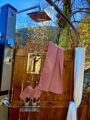 A wooden outdoor shower with a metal showerhead, a towel rack with towels, and a bottle of shampoo.