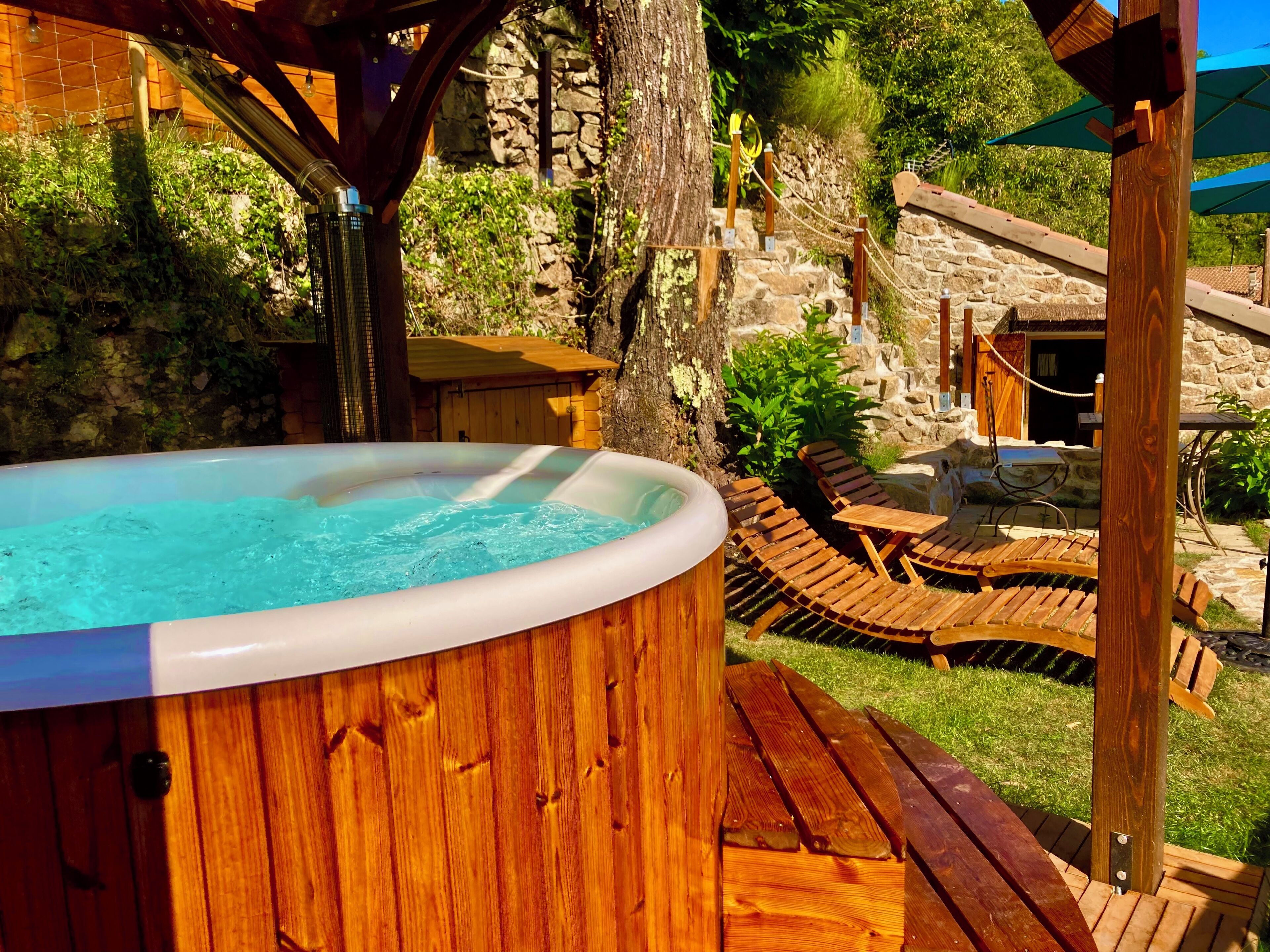 Un bain à remous installé sur une terrasse en bois, avec des chaises longues et un mur de pierre en arrière-plan.