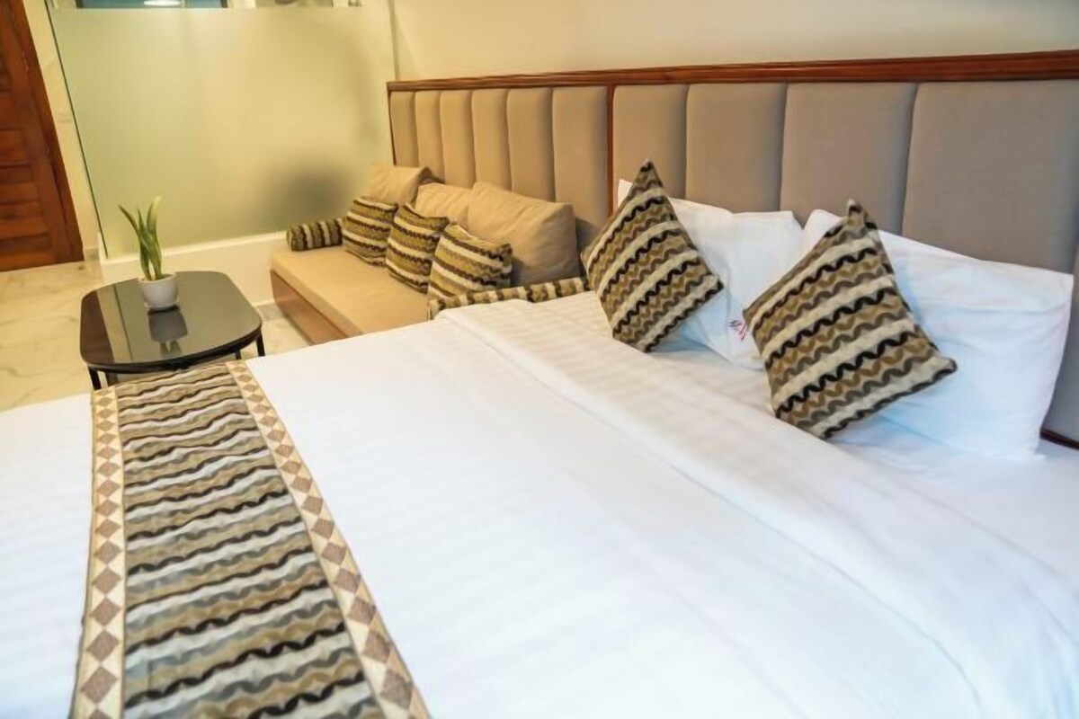 A neatly made bed with patterned pillows and a striped throw, a small round table with a plant, and a sofa with matching cushions.
