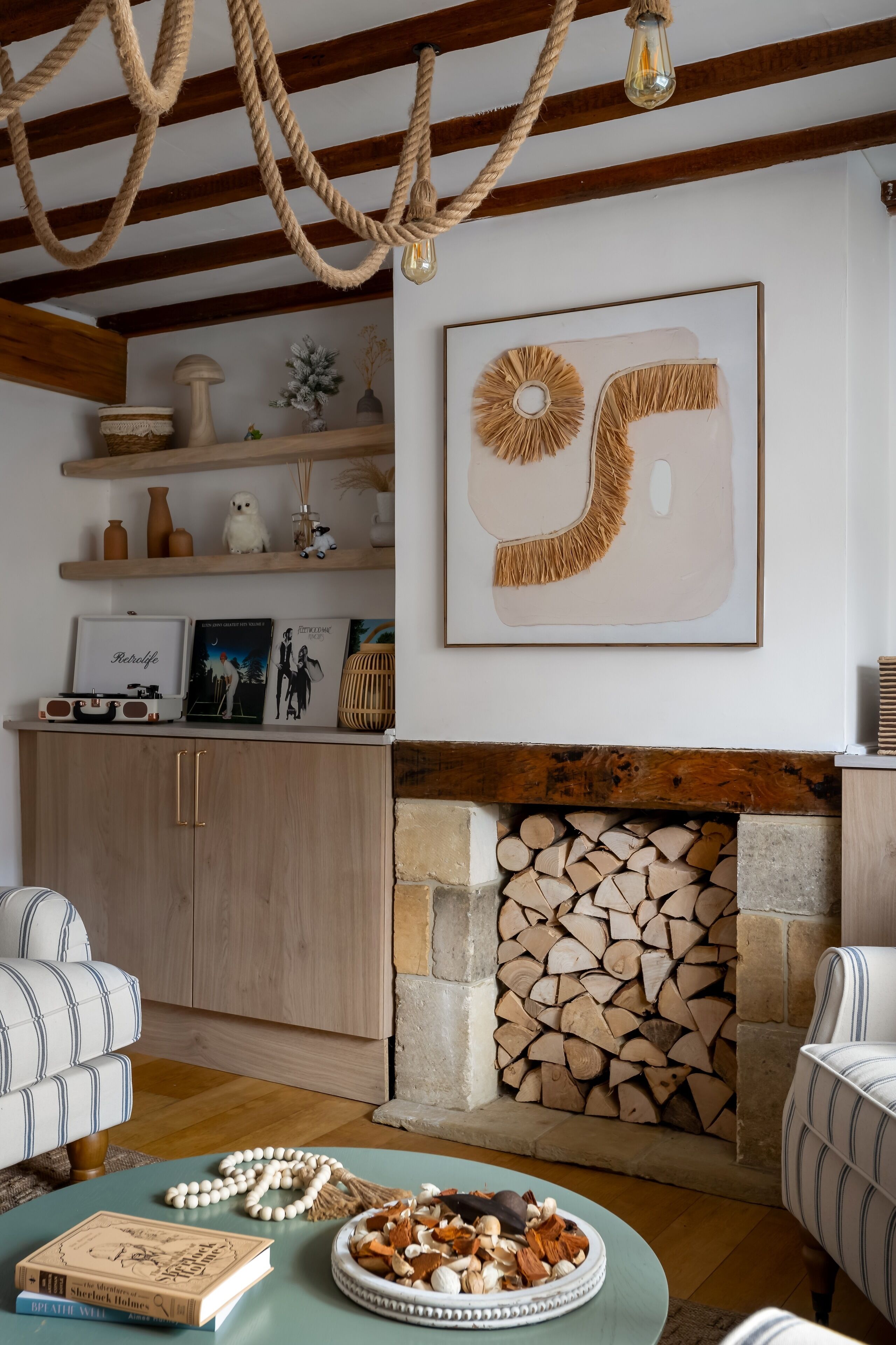 Un salon chaleureux avec une cheminée, des poutres en bois, un fauteuil à rayures et une table basse agrémentée de livres et d’objets décoratifs.