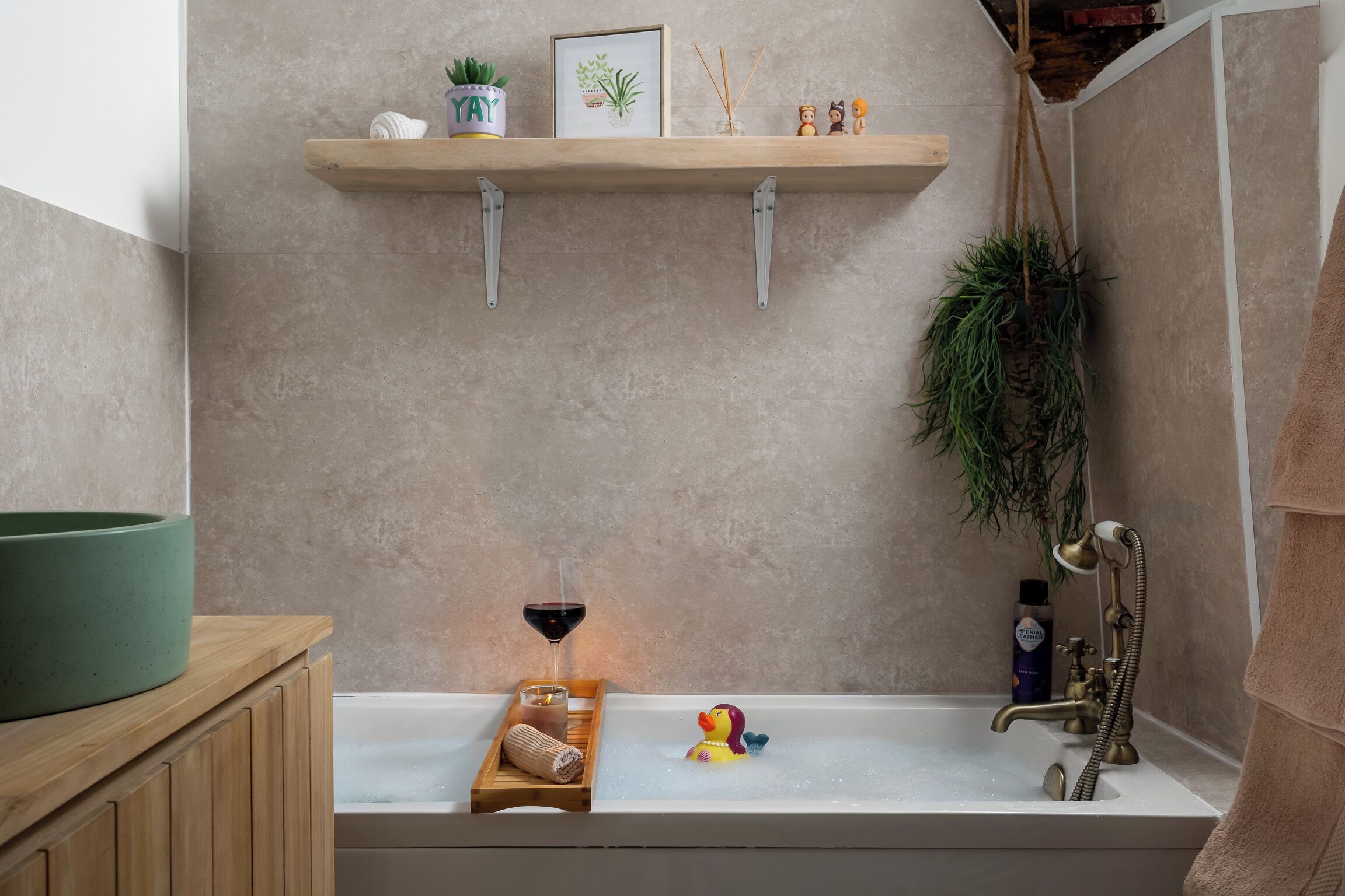 A bathroom with a bathtub, a shelf with plants and a framed picture, a glass of wine, a rubber duck, and a towel rack.