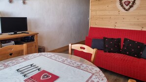 Ein gemütliches Zimmer mit einem Holztisch, einem Fernseher, einem roten Sofa mit gemusterten Kissen und einer herzförmigen Dekoration an der Wand.