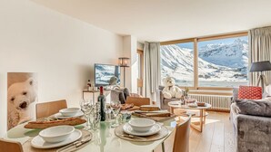 A dining area with a glass table set for four, a sofa, and a large window with a snowy mountain view.