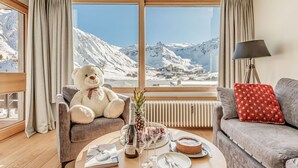 A cozy room with a large window, a plush sofa, a chair, a table with a book, a bottle, glasses, and a dessert.
