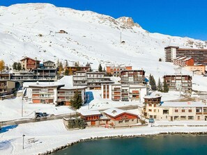 Un poble de muntanya cobert de neu, amb edificis moderns, un llac petit i un cel blau clar.