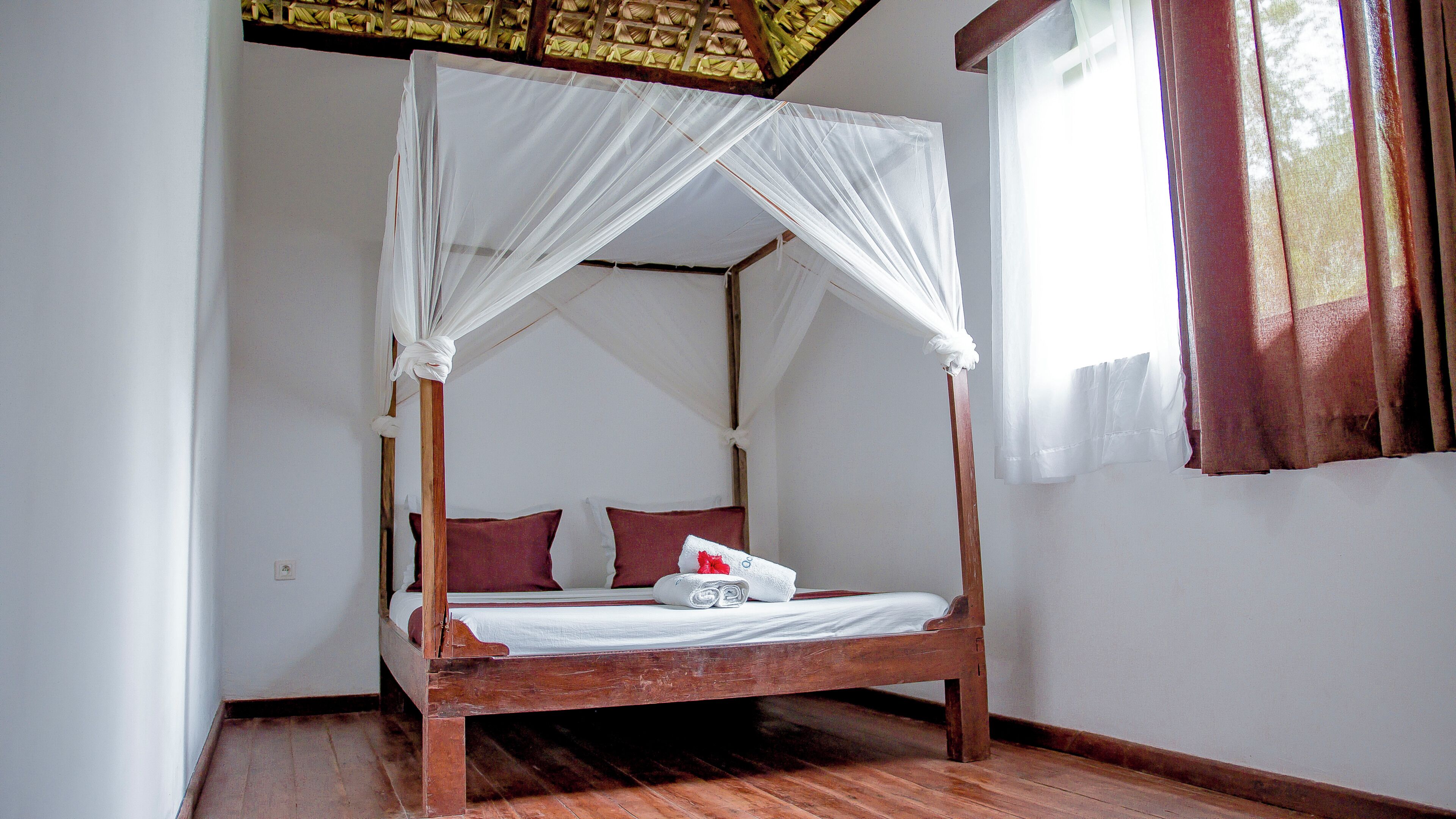 A wooden four-poster bed with white curtains, a red pillow, and a white towel on the bed.