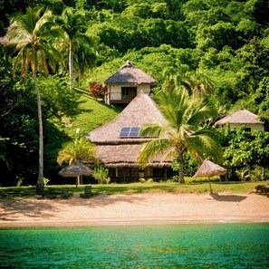 A tropical resort with thatched-roof buildings, palm trees, and a beach. - Hotel Ocean Beach (Nosy Be)